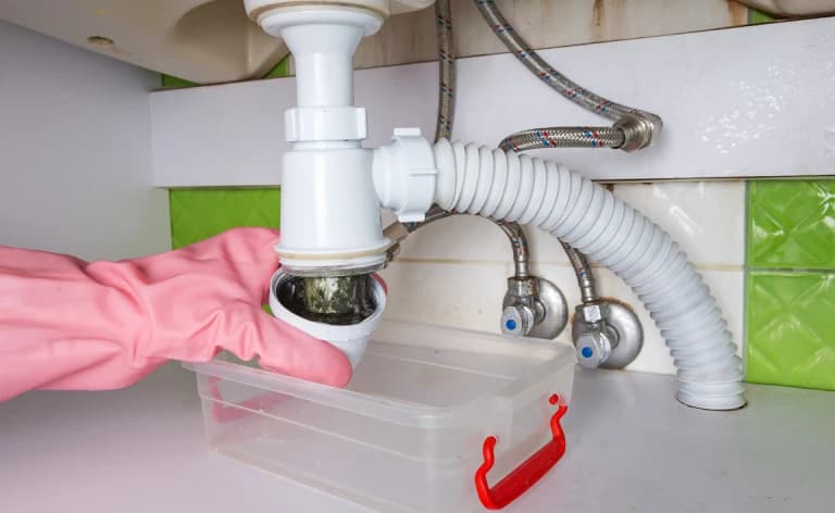 A pink-gloved hand unscrewing a sink trap over a plastic container.