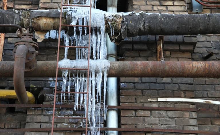 Thick ice hanging from exposed pipes beside a brick wall.
