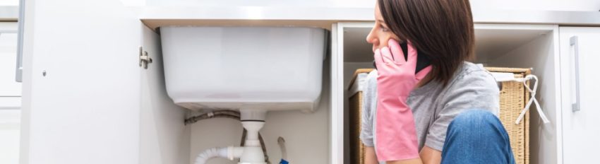 A woman calling a plumber while attempting to fix a leak under the kitchen sink